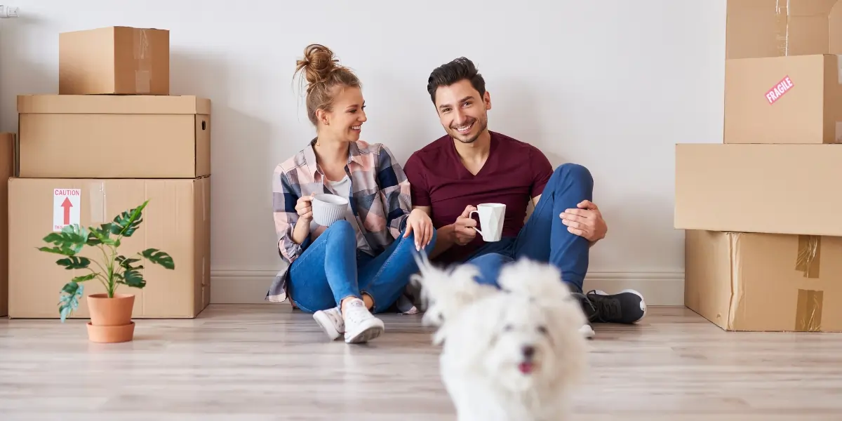 Two renters taking a coffee break on the floor among stacked moving boxes in their new apartment, with their small dog walking by